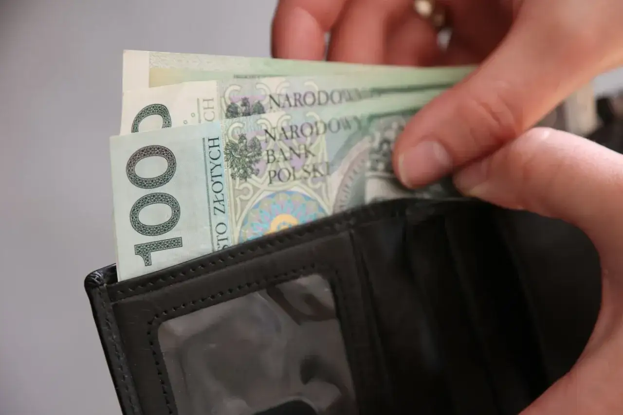 Ręce wkładają banknoty 100 zł do czarnego portfela. To może być dodatek za wysługę lat w budżetówce.