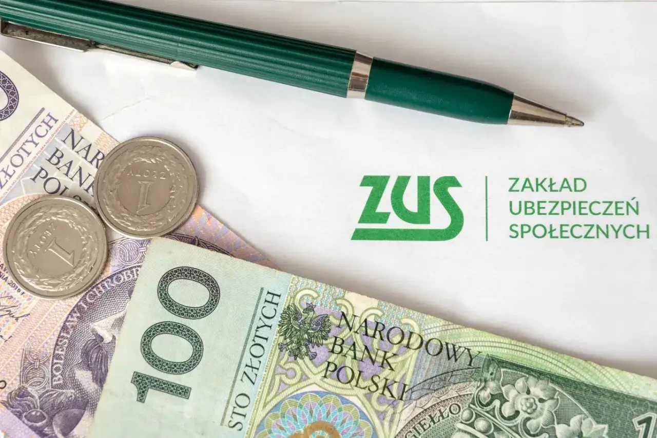 Pieniądze, monety i długopis na tle logo ZUS. Symbolizuje to wypłatę gwarantowaną ZUS.