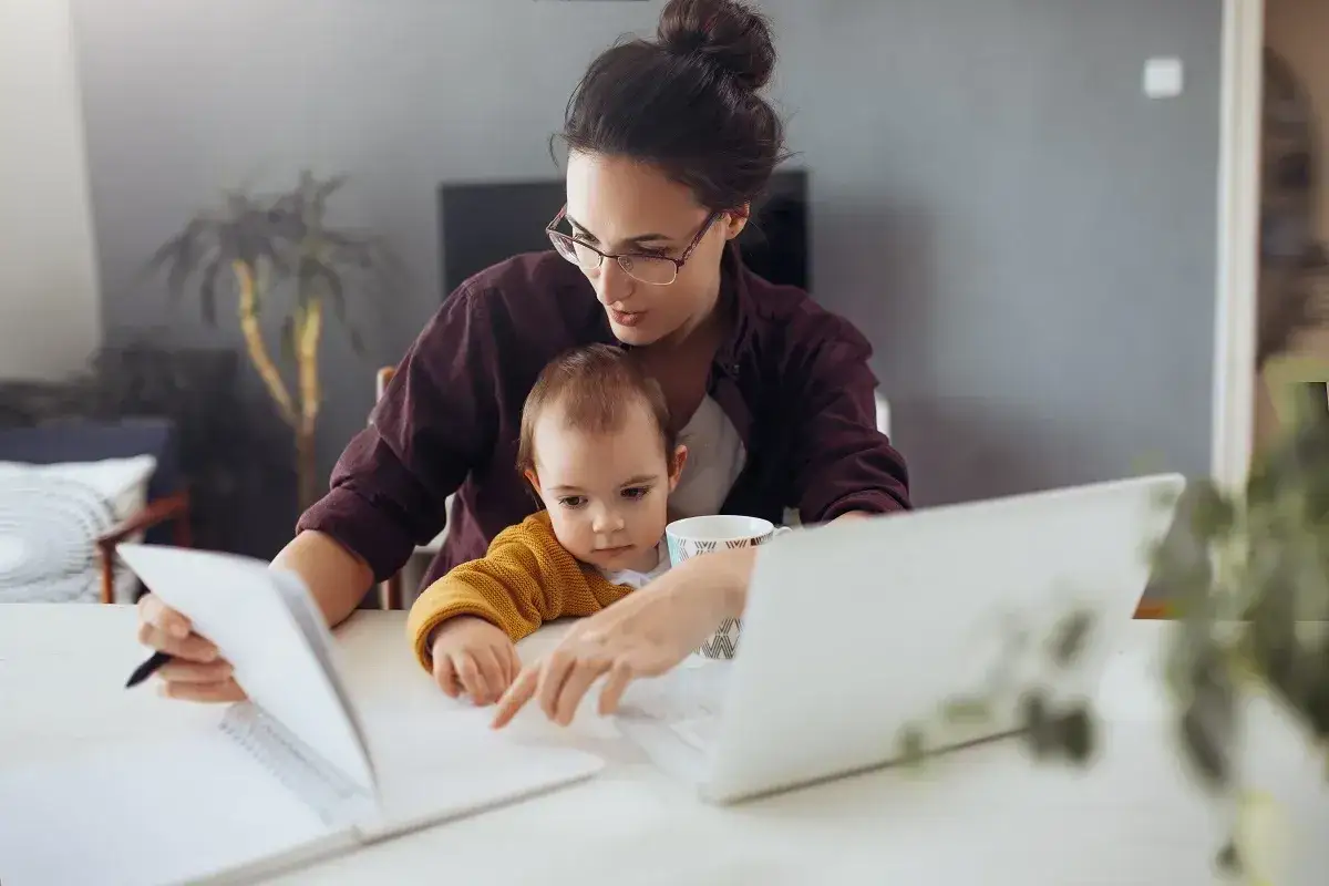 Mama z dzieckiem na kolanach pracuje przy laptopie, zastanawiając się, czy na macierzyńskim można pracować.