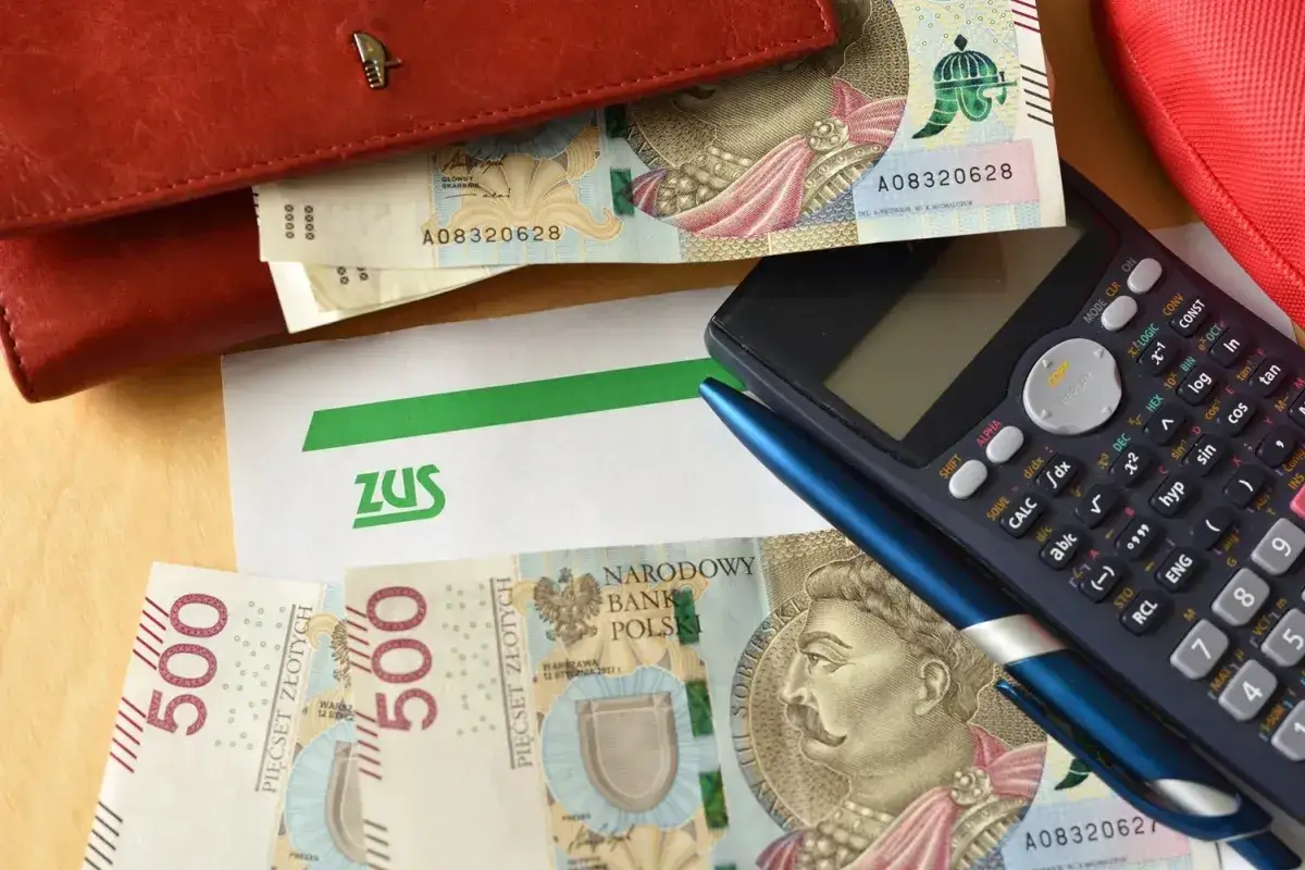 ZUS i pieniądze na stole. Obok kalkulator i długopis sugerują planowanie finansowe, być może związane z wypłatą gwarantowaną ZUS.