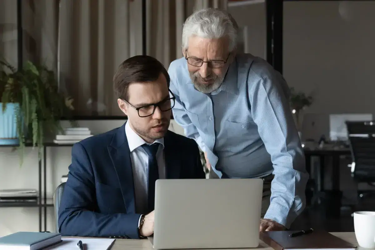 Dwóch mężczyzn analizuje dane na laptopie. Starszy pracownik tłumaczy młodszemu zasady naliczania dodatku za wysługę lat w budżetówce.