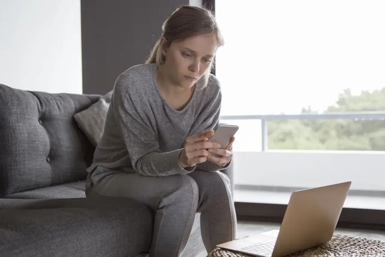 Kobieta w dresie siedzi na kanapie, przeglądając telefon. Obok leży laptop, sugerując pracę zdalną lub umowa zlecenie.