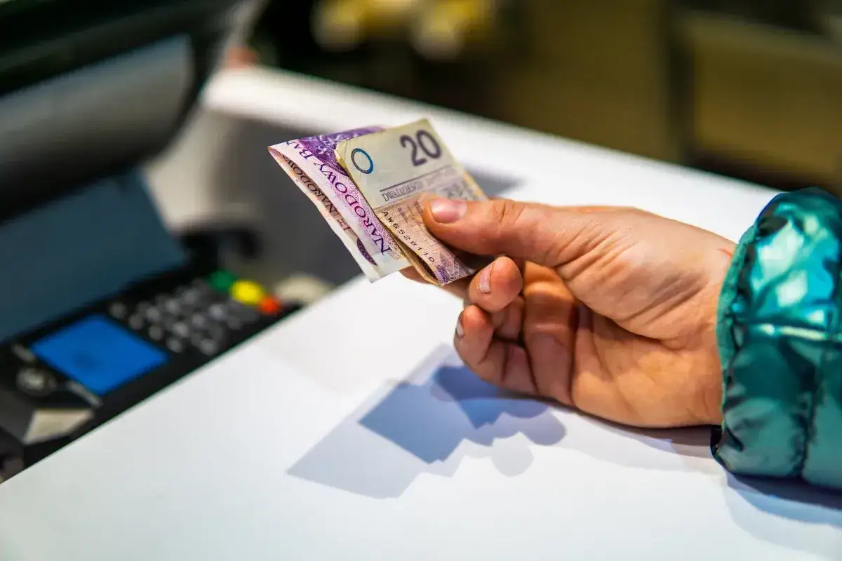 Dłoń trzyma banknoty, przygotowując się do zapłaty. W tle widać terminal płatniczy, sugerujący transakcję i zwolnienie z kasy fiskalnej.